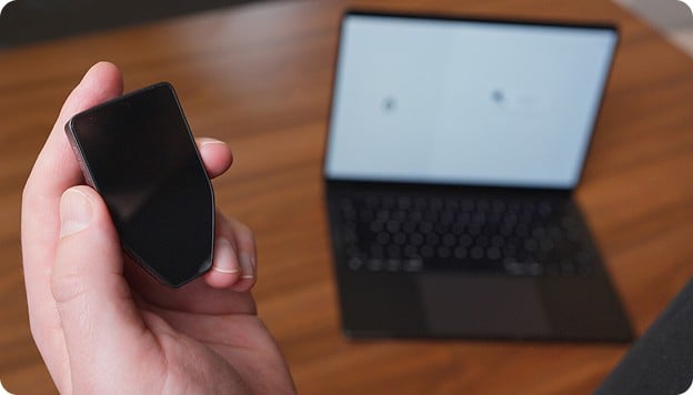 Hand holding a small black device in front of an open, blurred laptop on a wooden desk.